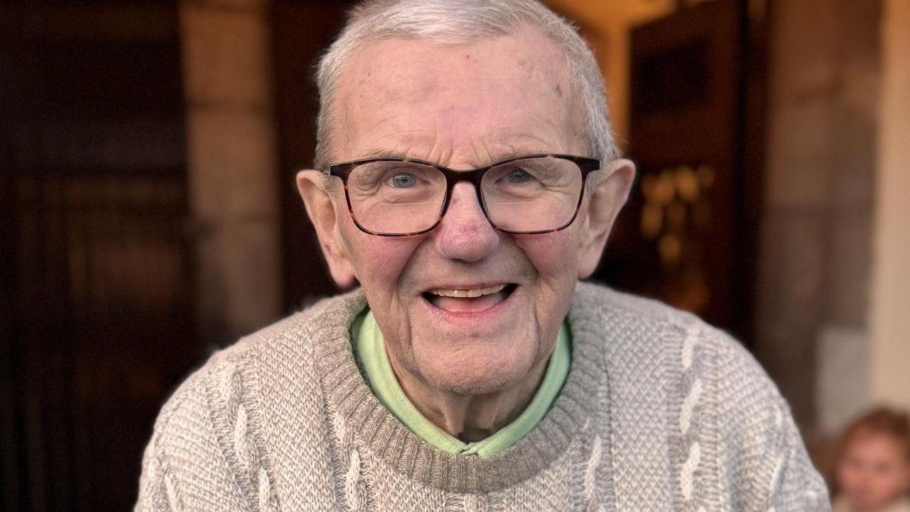 Gerry, a man with white hair and large reading glasses wears a grey jumper and is pictured in a church doorway.