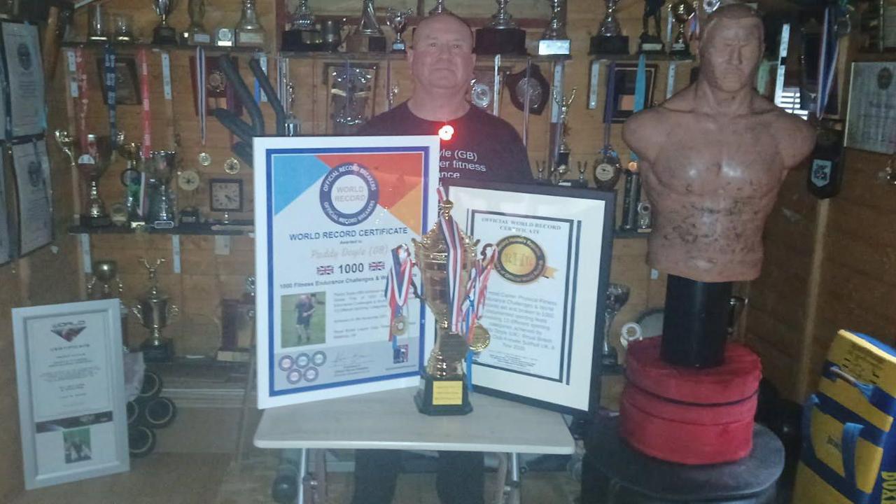 A bald man in a black top standing behind two large framed awards and a gold trophy with medals hanging off it. There are dozens of trophies and awards on shelves in the background, and a boxing torso equipment next to him.