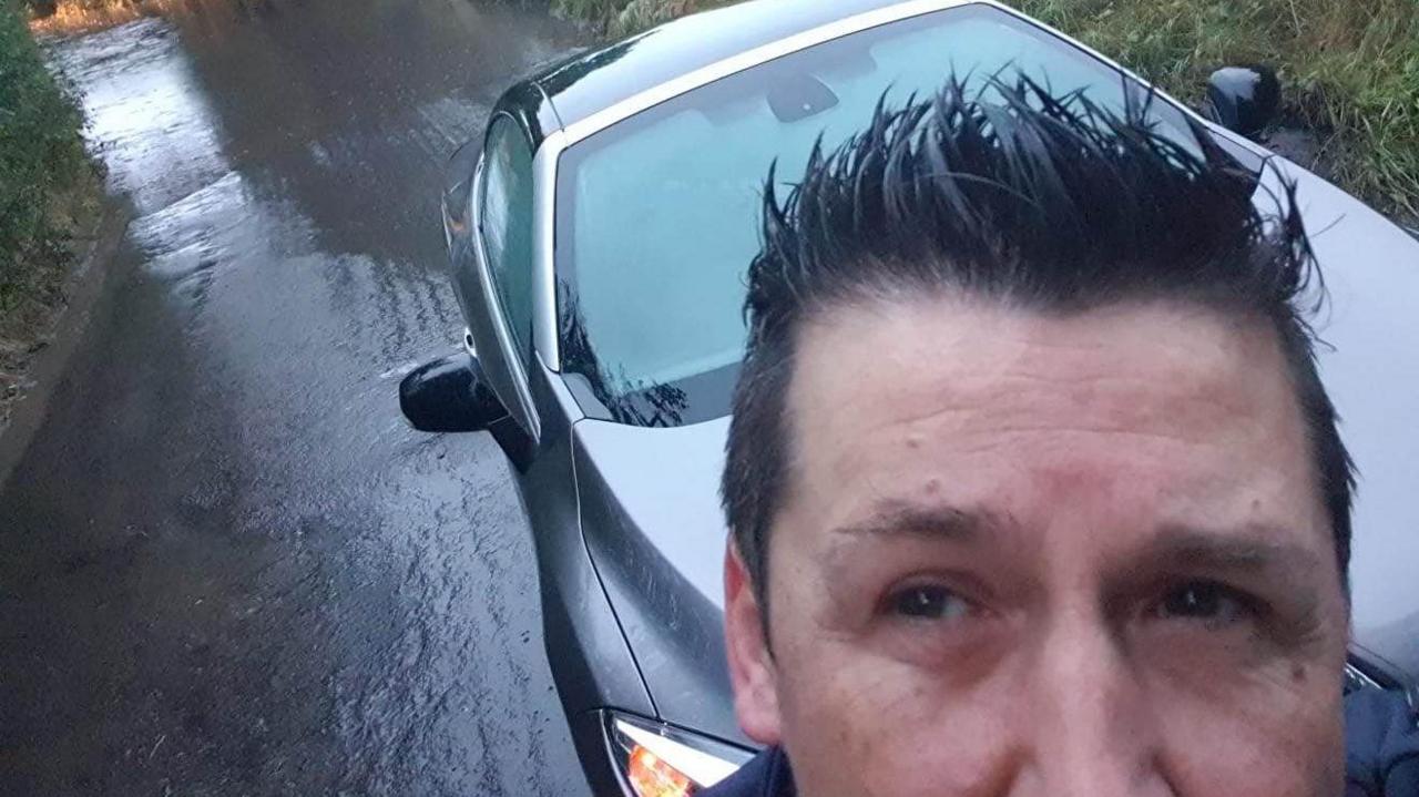 A man with dark spikey hair and a grey car is parked behind him. He is showing a pool of standing floodwater on a muddy narrow country road.