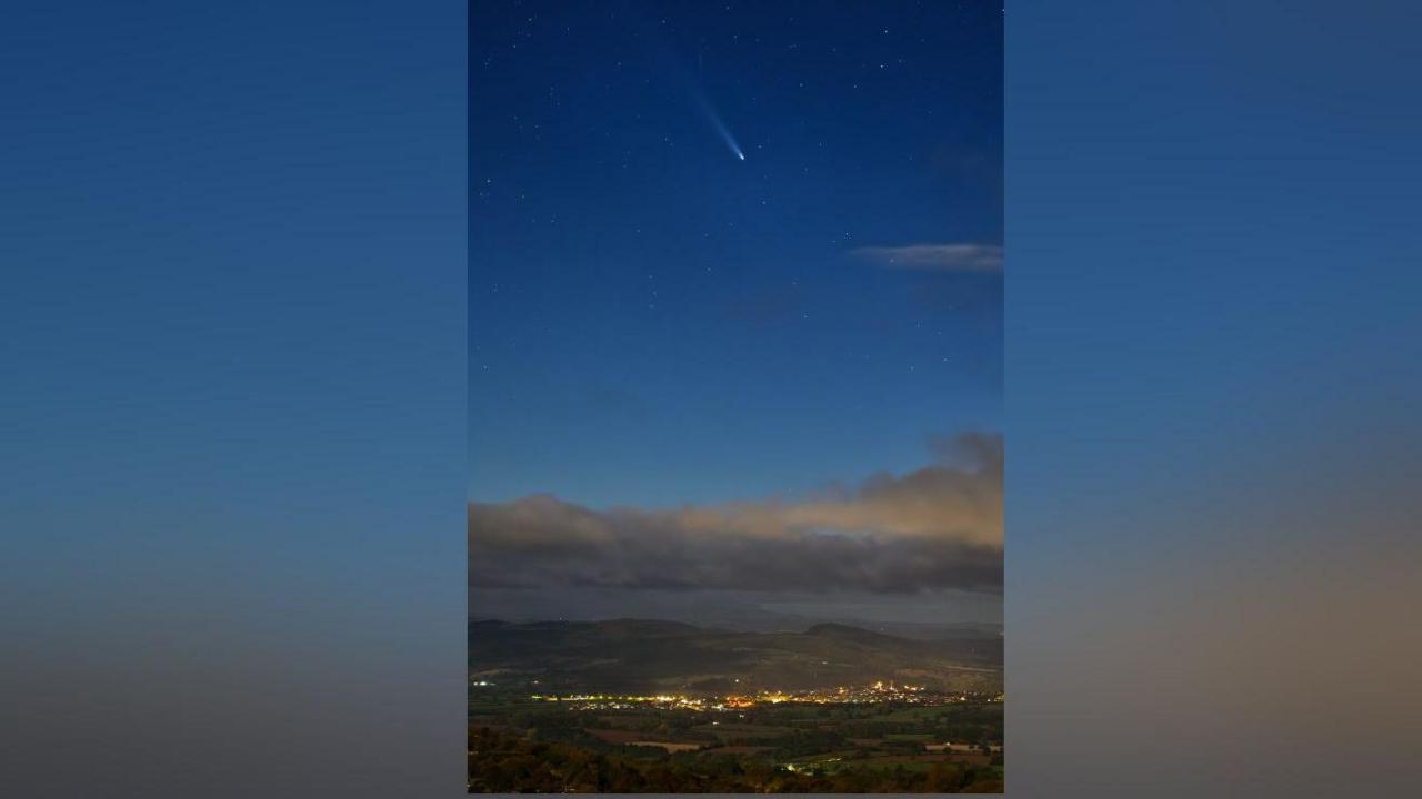 A comet in a dark blue sky above the lights from homes on the ground below and some grey cloud