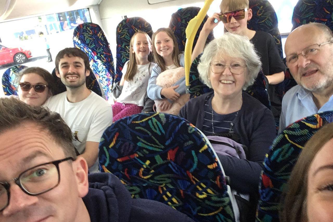 A group of nine people of various ages are smiling whilst posing for a photo. They appear to be on a bus. There are three children and six adults.