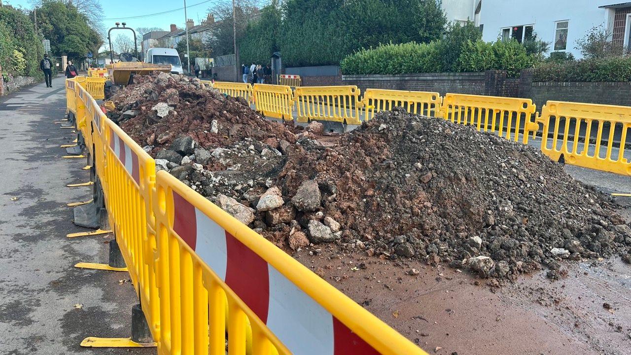 Plastic yellow fencing with red and white strips around a section of Heavitree Road that has been dug up.