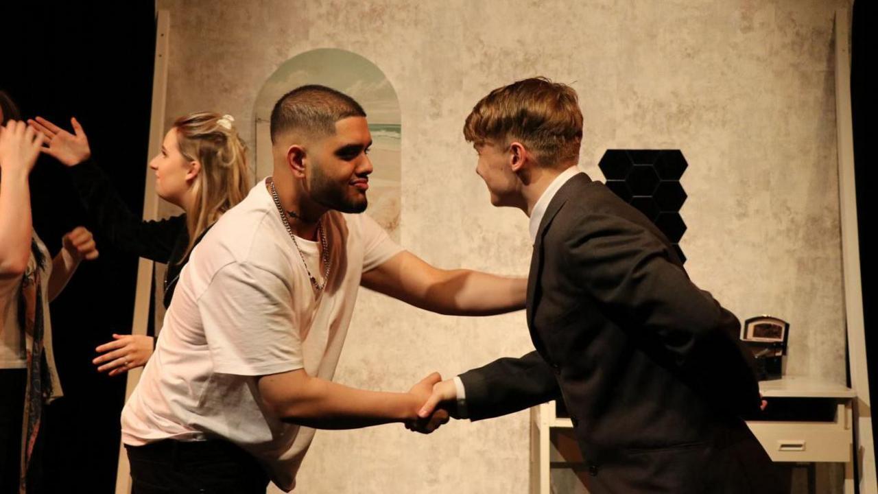 A man with short dark hair and a beard in a white t-shirt shaking the hand of a man with blonde hair in a dark suit.