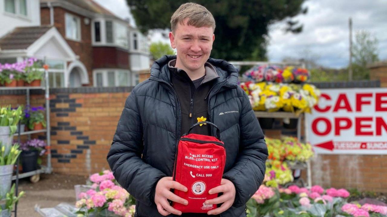 Bobby-Ray has brown hair and is wearing a dark coat. He is holding a red bag that contains a bleed kit and is standing in front of rows of flowers. There is a large white sign with red writing, that reads cafe open, behind him.