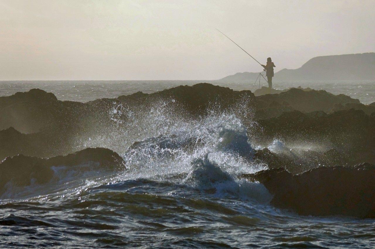 Huge waves in the foreground in front of a rocky outcrop which a man is standing on makes it look like he is fishing in a treacherous situation.