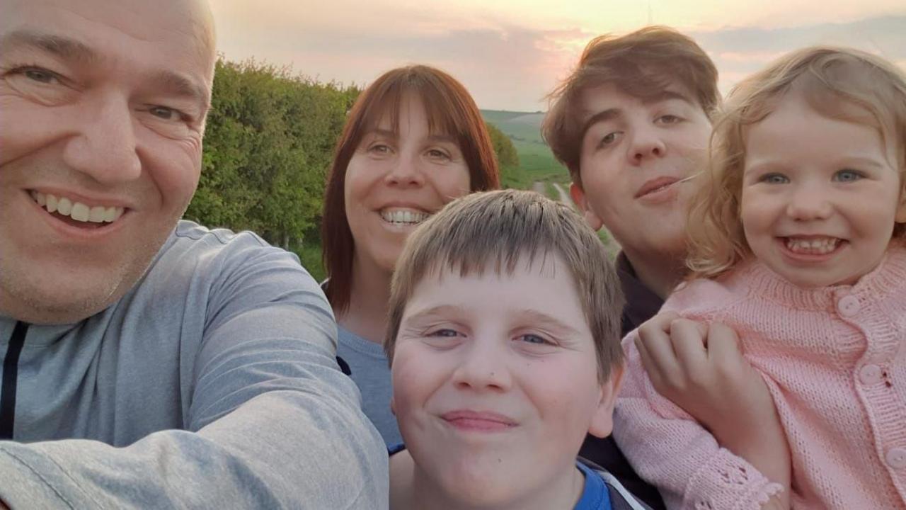 Three children, two boys and a little girl being held in the air by her brother smile at the camera. Serena is in the middle of the family group with her husband Mark to the side of the image. The whole family is huddled together with a sunset behind them, smiling.