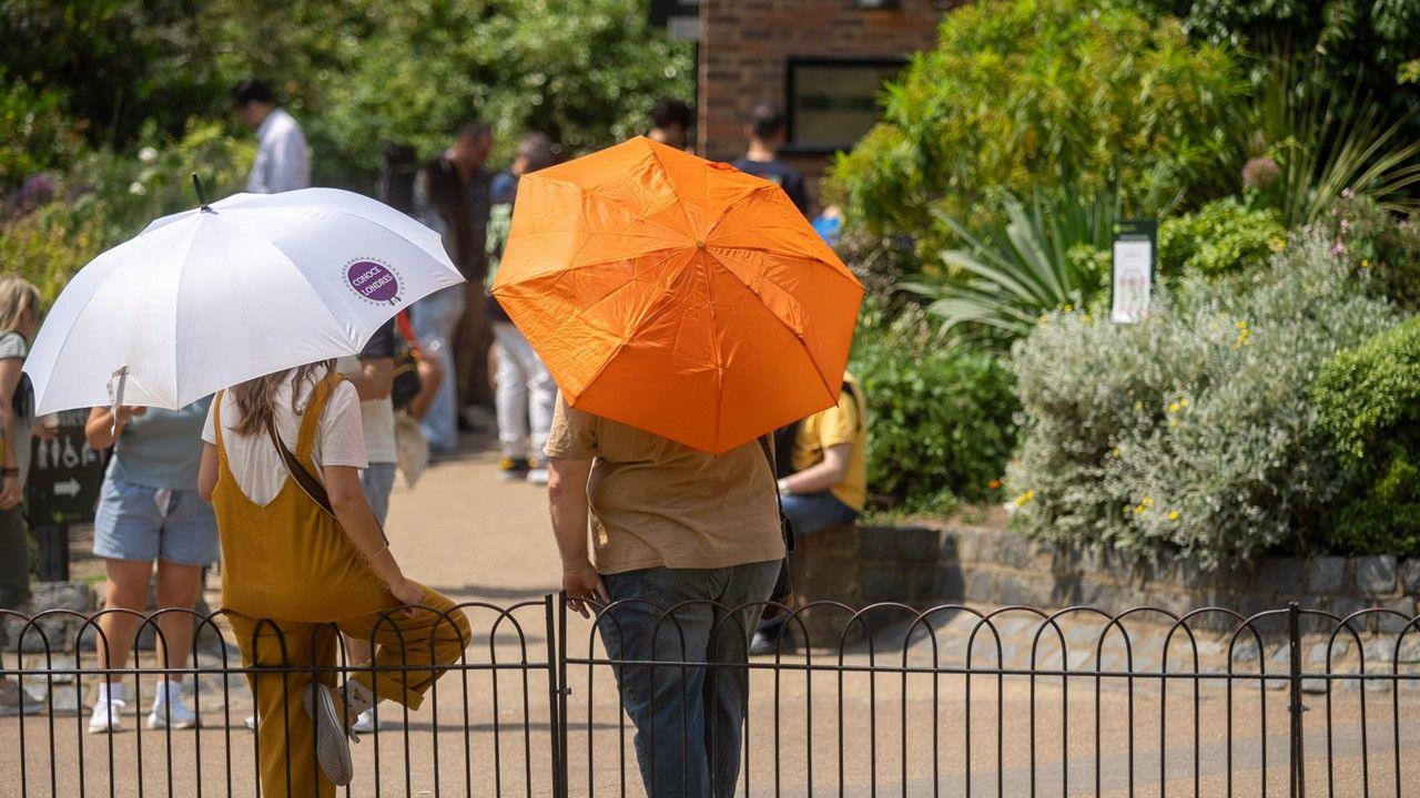 A woman and a man rest on railings with umbrellas up to shelter them from the sun. They are in a part and you can see shrubs and other people in the the background. 