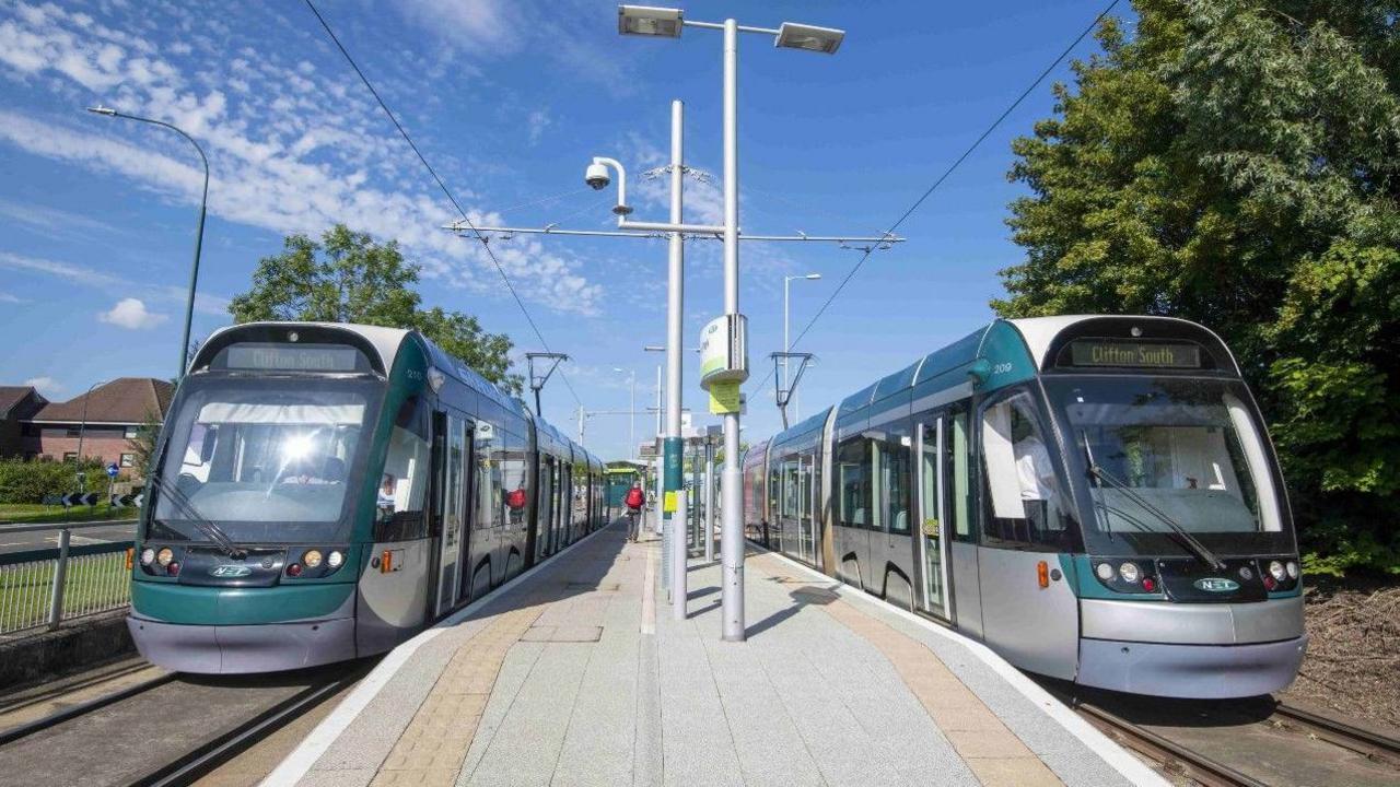 Two green and silver Nottingham trams pulled up at a stop. They are sitting either side of a raised concrete area for passengers to board from.