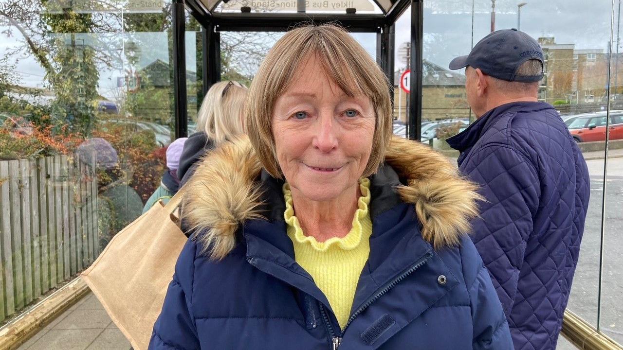 A woman with a short, dark blonde bob hairstyle wearing a blue coat with fury collar and a yellow jumper underneath standing in a bus stop. 