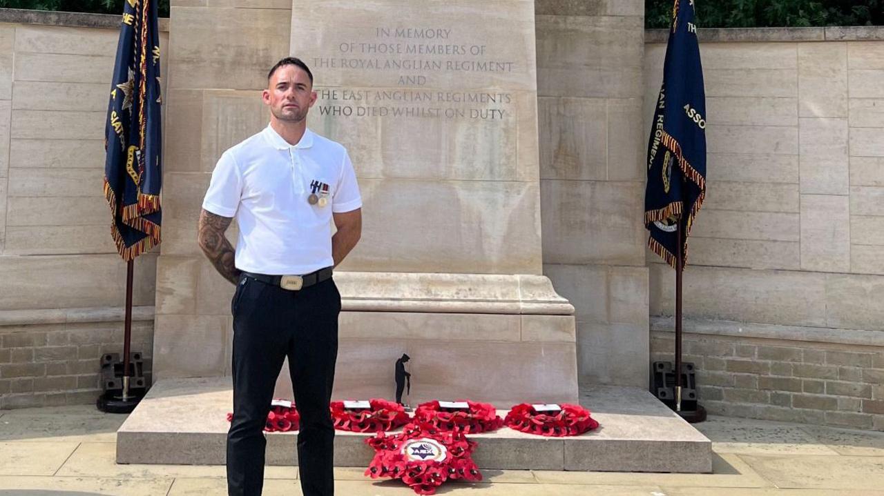 Rhys Thurtell wearing a white shirt and black trousers standing in front of a cenotaph and five read wreaths of poppies 