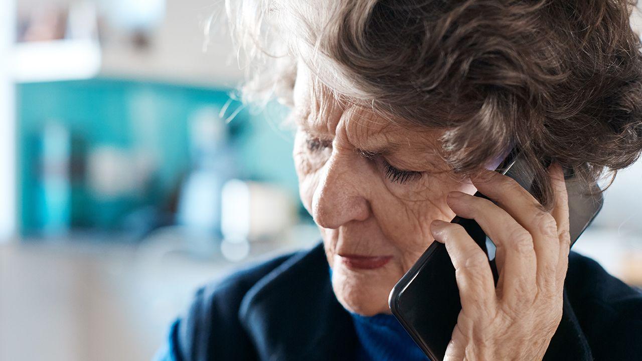 An elderly woman holds a phone to her ear while making a call. She looks concerned, with her eyes looking downwards, and her kitchen can be seen out of focus in the background.