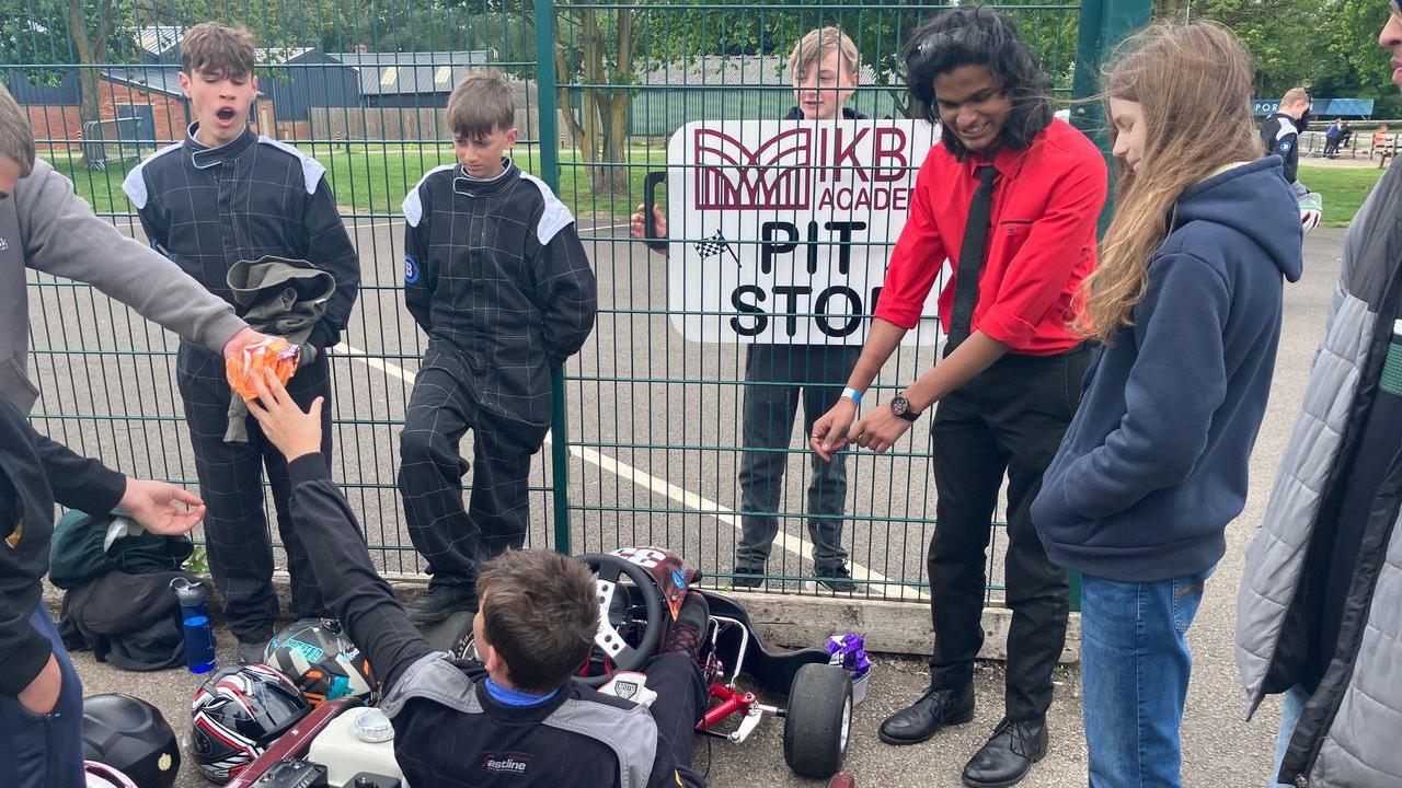 A group of young people in motorsport padded overalls stand over another young person in a small motorised go-kart