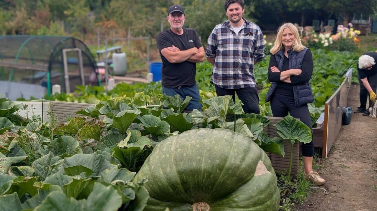 Two men and a woman posing infront of a giant squash in an allotment. 