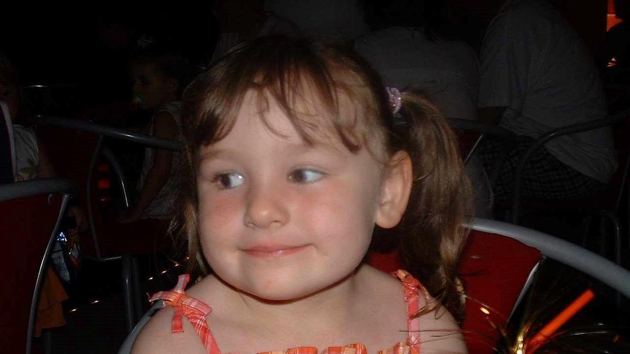 Rachel as a young girl with long brown hair with a fringe in pigtails with pink hair bobbles. She is smiling and wearing an orange gingham dress. 