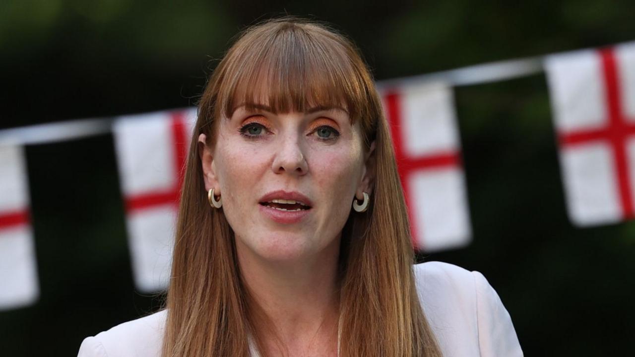 A red haired woman with a fringe and gold hoop earrings is speaking, standing in front of St George cross bunting.