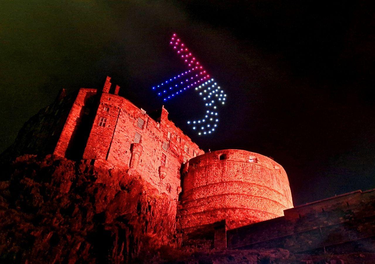 Edinburgh Castle, shot from below, covered in red light, with a drones flying above in the shape of the number 75