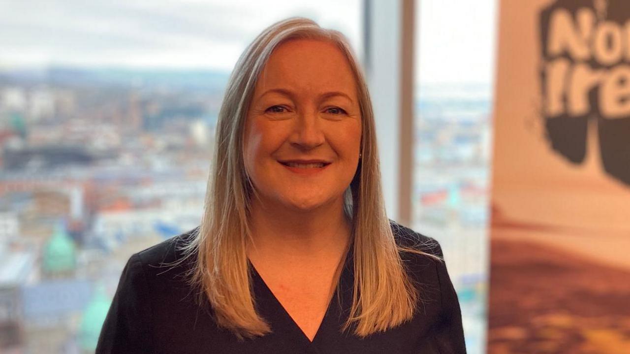 A blonde woman wearing a dark shirt smiles at the camera. Beside here is a sign which says Northern Ireland