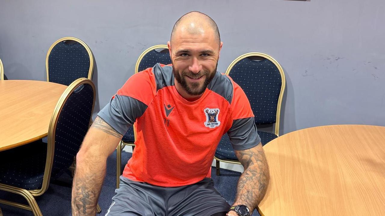 A man in a bright red football shirt with grey panels on the sleeves and shoulders sits at a round table in a conference room. He's smiling, casually leaning one of his heavily tattooed arms on the table next to him.