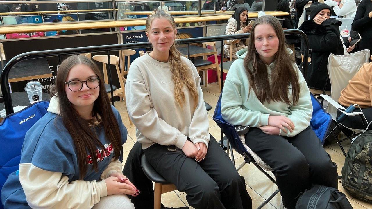 Three girls are sitting on chairs and blue camping chairs smiling. They are all wearing jumpers and trousers. 