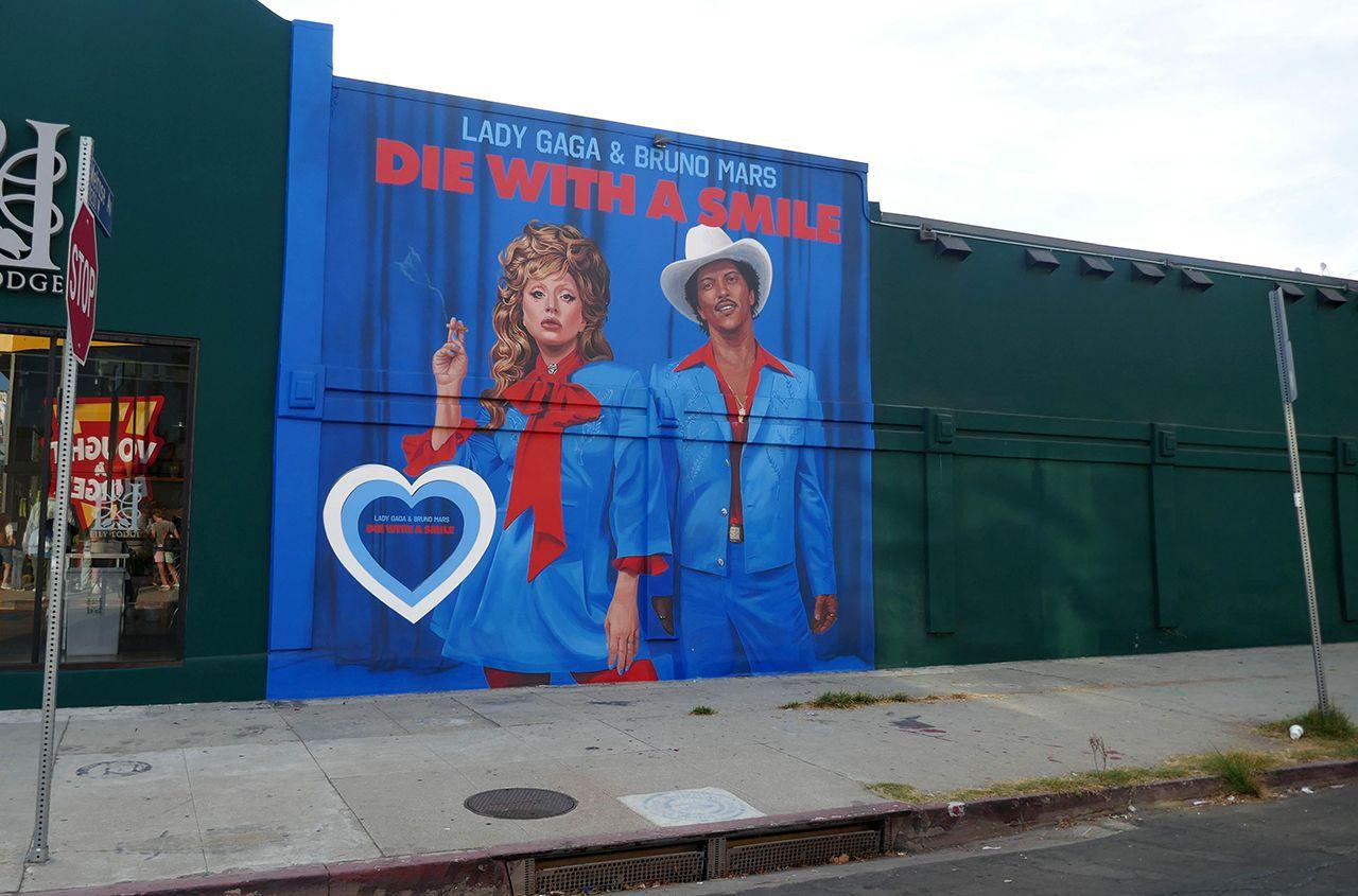 An art mural billboard depicting the album artwork for Lady Gaga and Bruno Mars' new single Die With A Smile. In the artwork, they both wear vintage looking blue suits with red collars, against a blue backdrop. Also in frame is a stop sign, a pavement, road and sky.