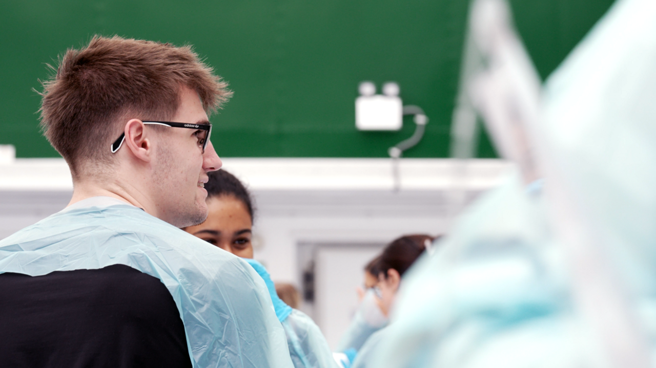 A male student with glasses and brown hair wearing a pale blue plastic apron sits in front of a girl with black hair in a university lecture. 