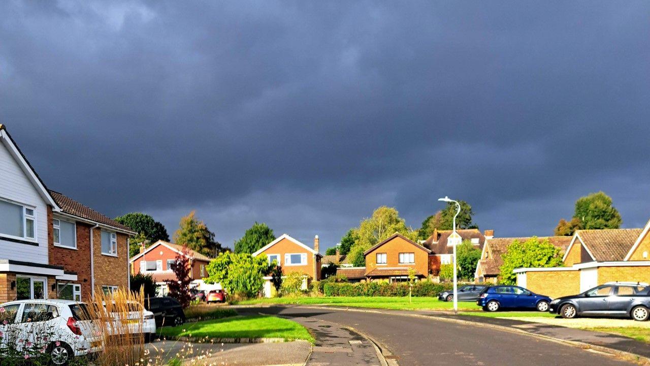 Dark grey clouds fill the sky over a curved suburban road with houses and cars parked in front