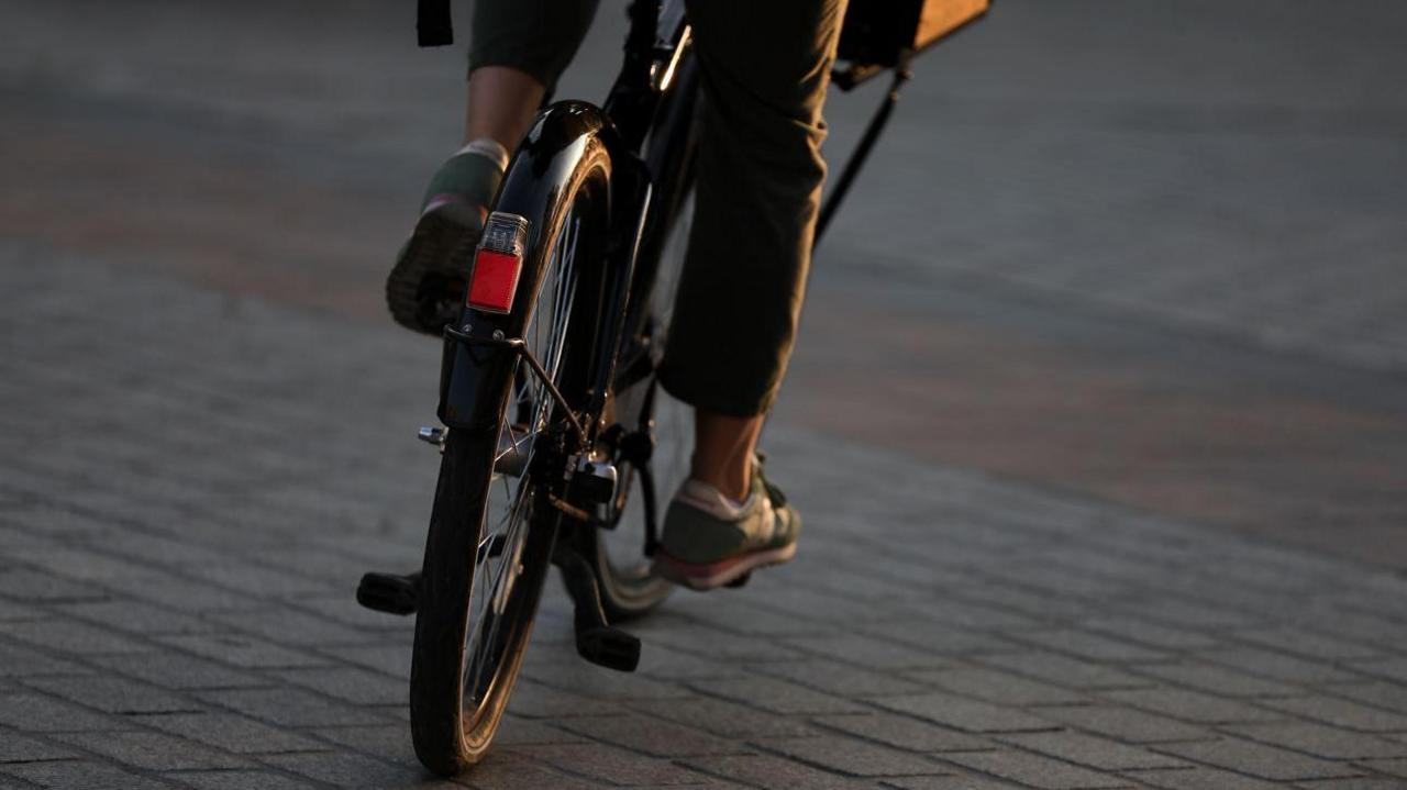 Close up of legs wearing track suit and trainers riding a bicycle across paving stones away from the camera  