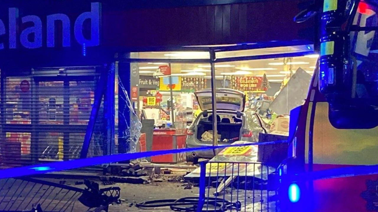 A car with the boot up inside an Icelend store. In a photo taken from the street, part of a main Iceland sign outside the store is visible. Signs inside include Fruit & Veg and Frozen.