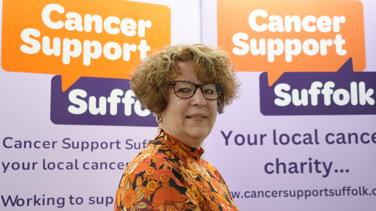 A head and shoulders image of Elizabeth Love-Mott. She is wearing a colourful orange, yellow and black top and glasses. She is standing in front of a back drop featuring the Cancer Support Suffolk logo. 