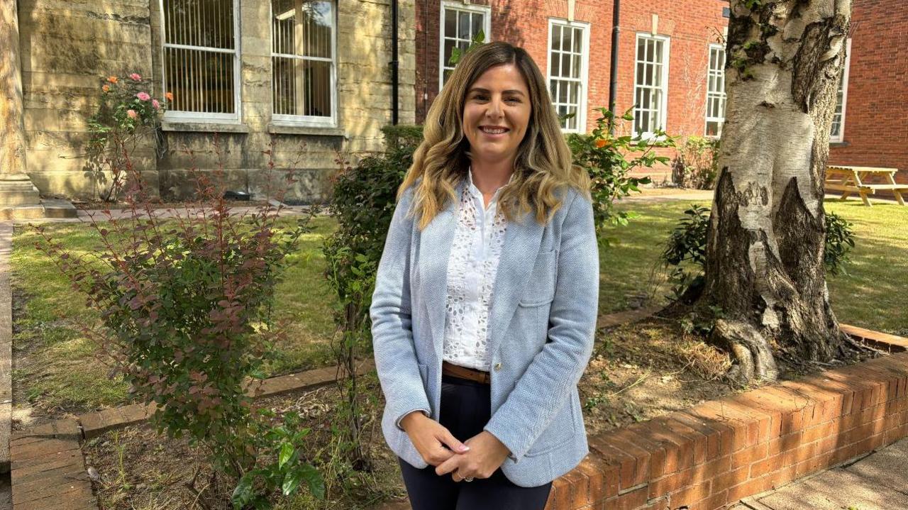 Natalie wears a light blue tweed jacket and has long midbrown curled hair. She stands in front of two roses and a tree on a sunny day in front of the council buildings.