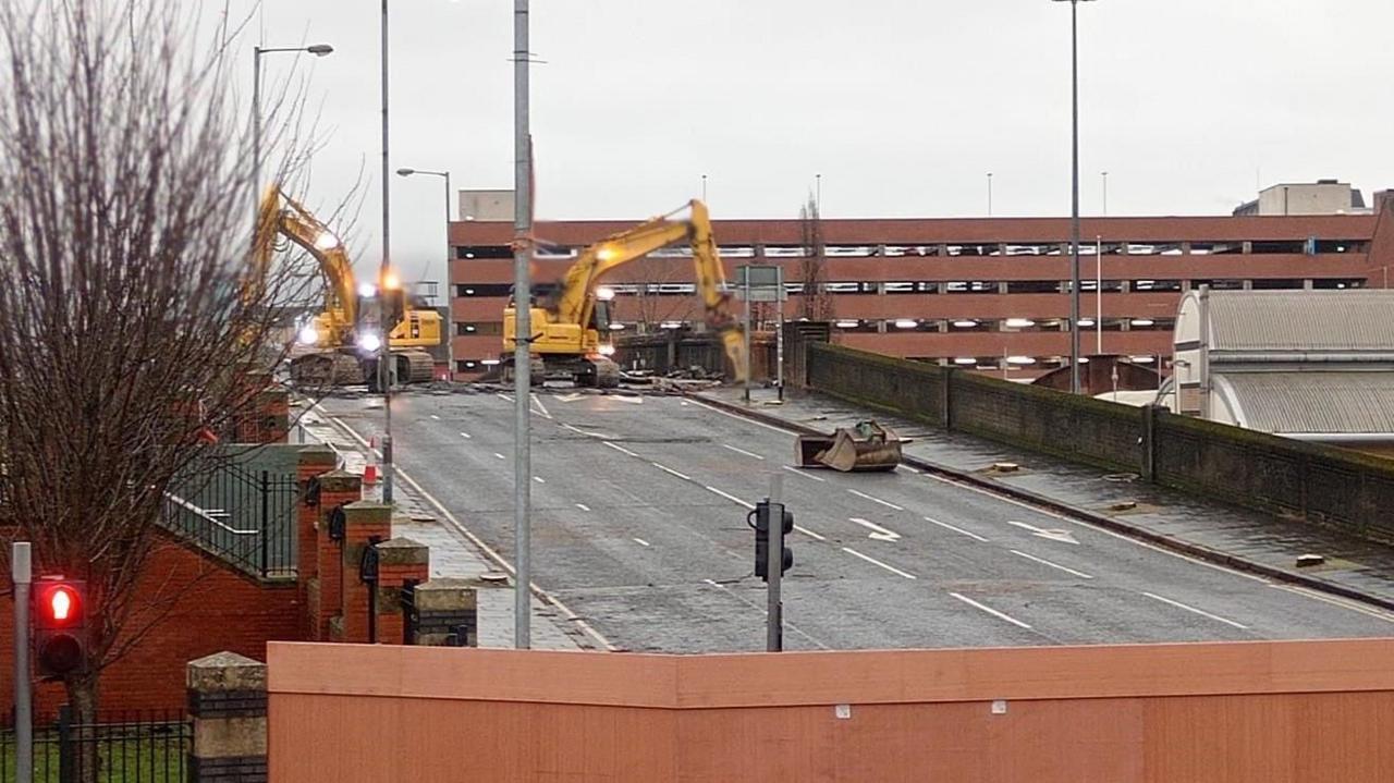 The Boyne Bridge in Belfast with yellow diggers working. There are trees on the left and buildings in the distance.