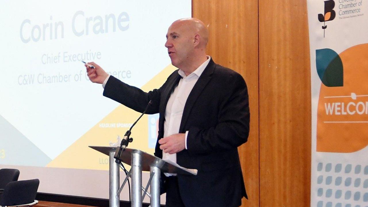 A man with a dark jacket and white shirt is talking to an audience. His right arm is outstretched and holding a pen. His name is on the screen behind him.