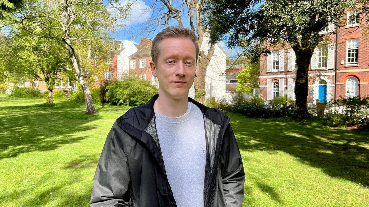 Ryan Hill in a grey top and a black jacket, standing in Southernhay Gardens with trees and houses behind him.