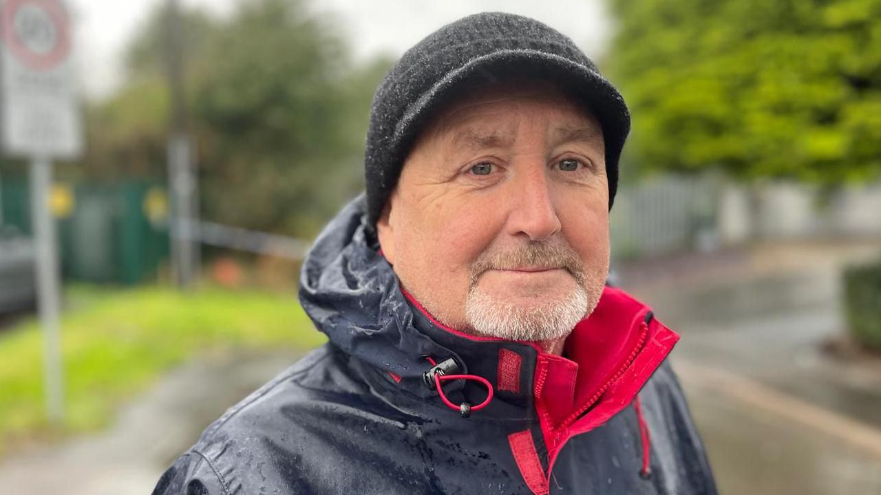 A man is wearing a wet blue raincoat with a red inside trim. He has a beard and is wearing a black woolly hat which is wet with the rain