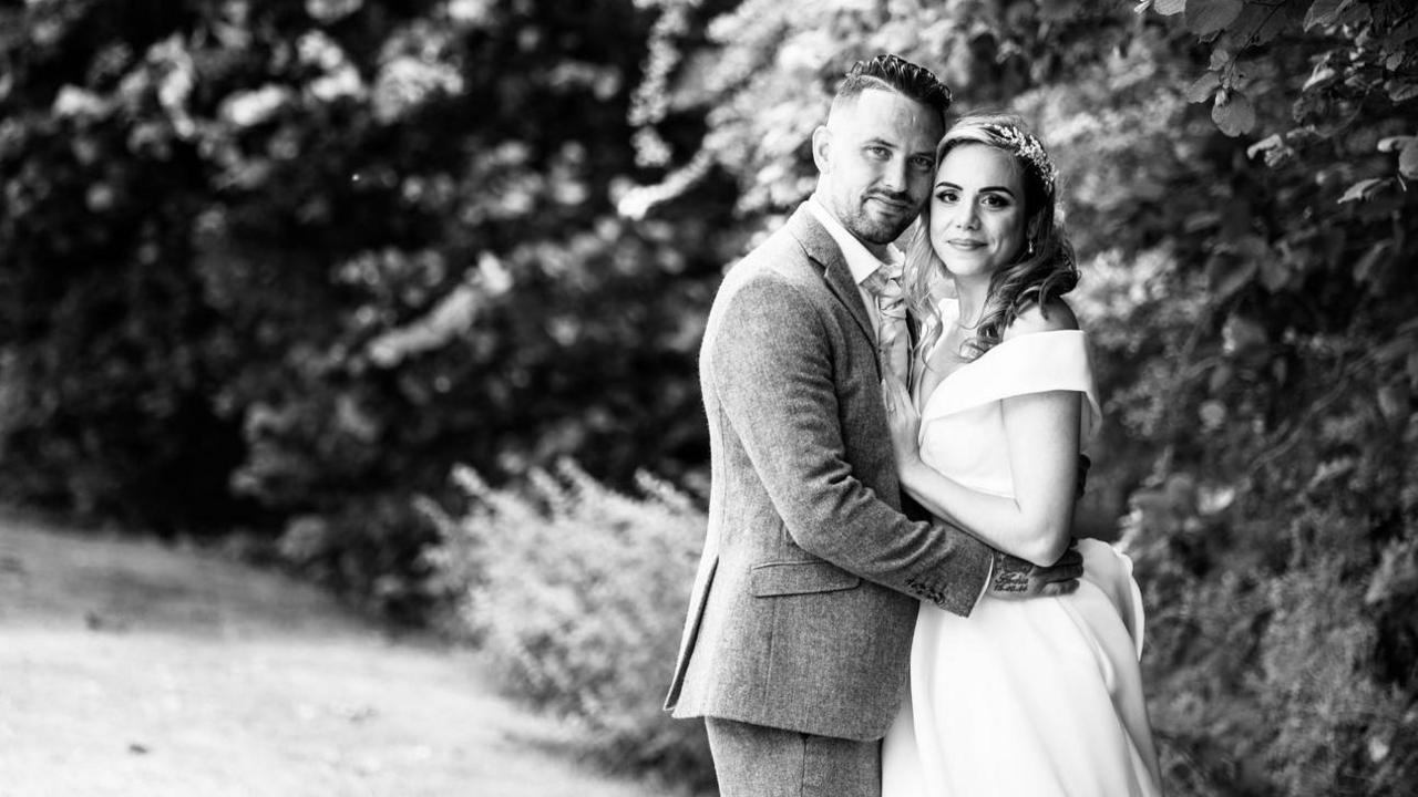 Jason and Mimi pose for a black and white wedding photo next to some trees.