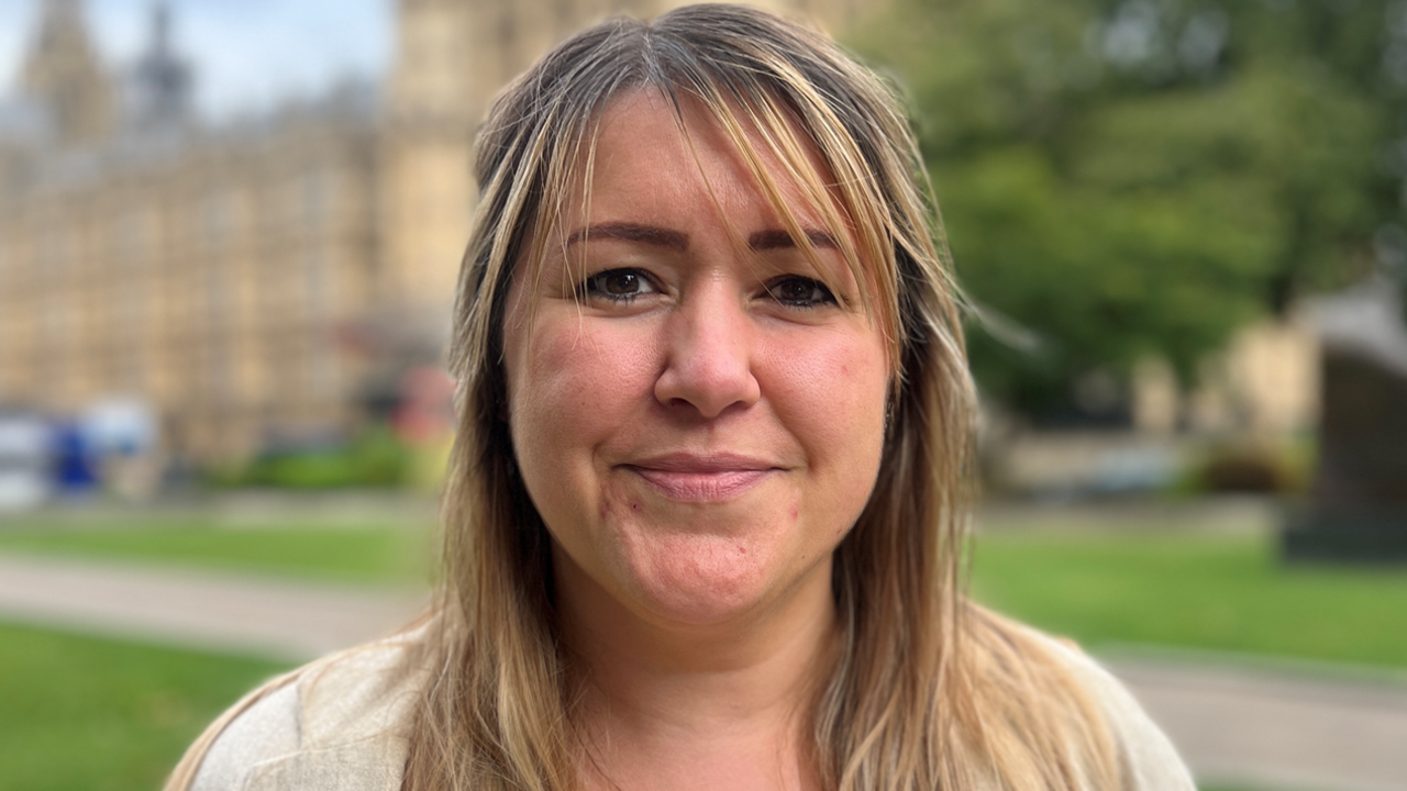 A woman with long brown hair and a fringe, wearing a pale top and standing outside the Houses of Parliament