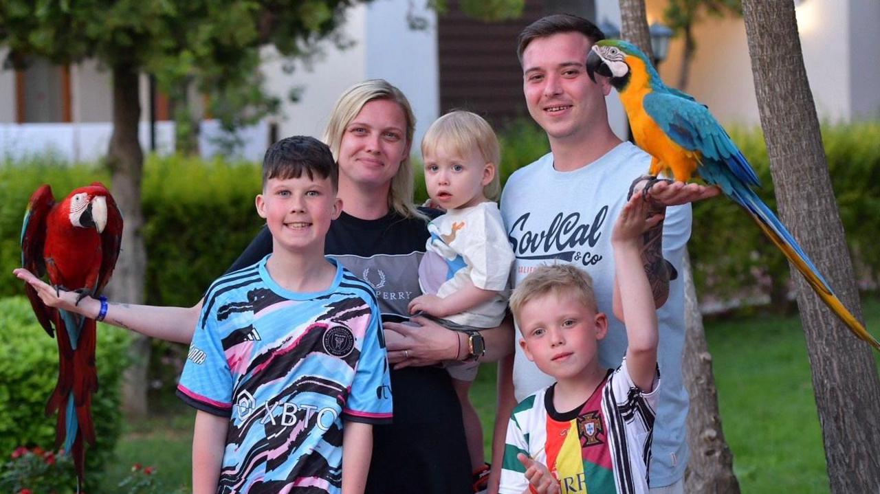 The family together on holiday. Harley and Louie are wearing a sports top. Their parents are in the background. Sophie is holding a baby. There are two birds. 