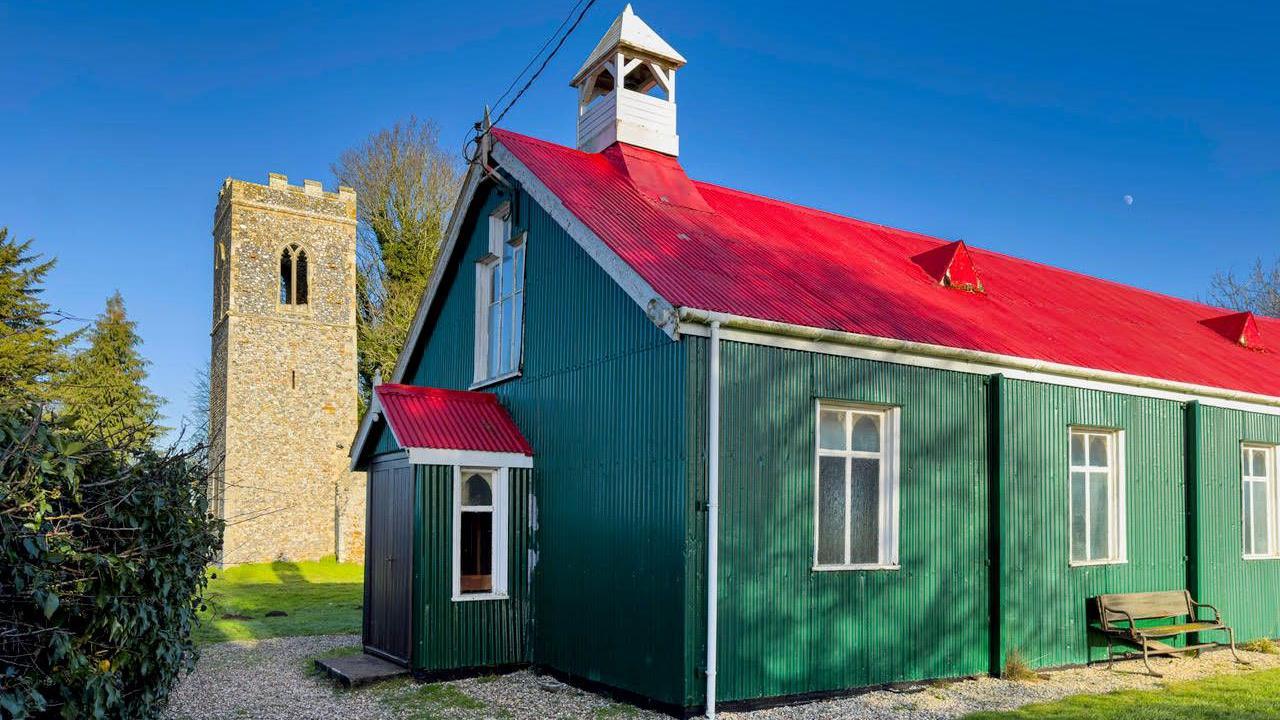 A green building with a red roof that has been built out of corrugated iron. It sits in front of a large stone church.