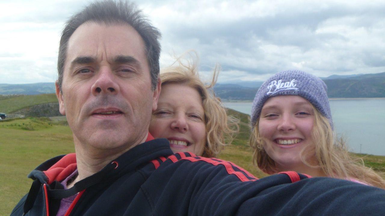 A picture of a man, woman and a teenage girl smiling at the camera standing on top of a hill. Mountains and the sea are in the background.