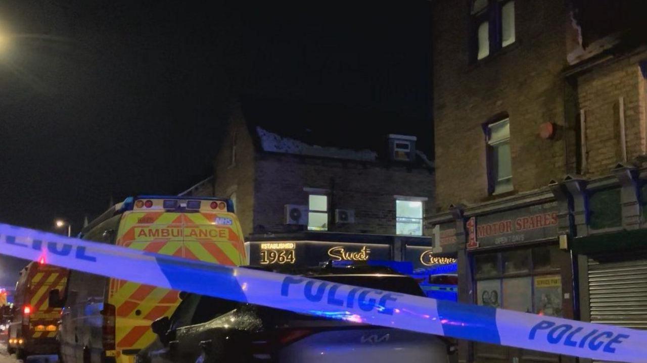 Police tape cordons off part of a street. Two ambulances can be seen parked on the street.