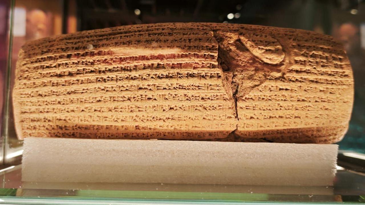 A replica of the Cyrus Cylinder in the Parsi museum covered by cuneiform script, displayed in a glass case.