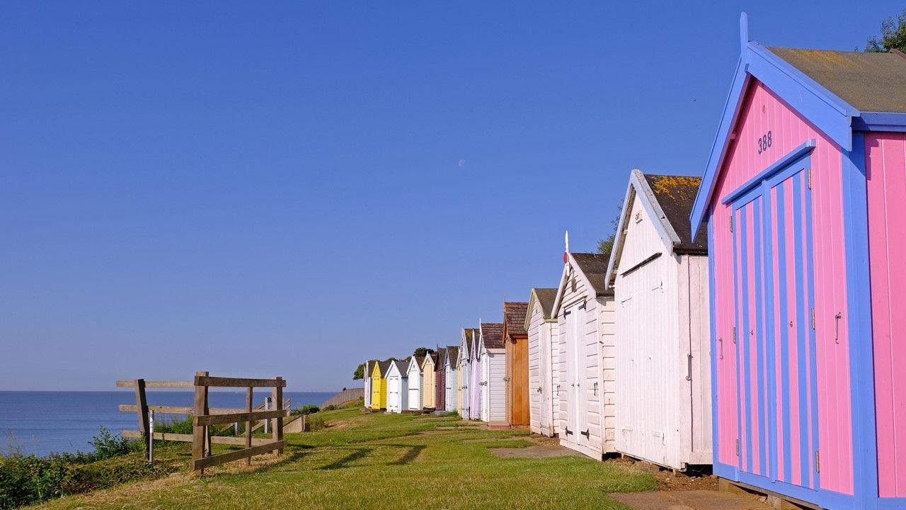 A row of multi-coloured beach huts on the right with some grass and a view over looking blue sea and bright blue sky