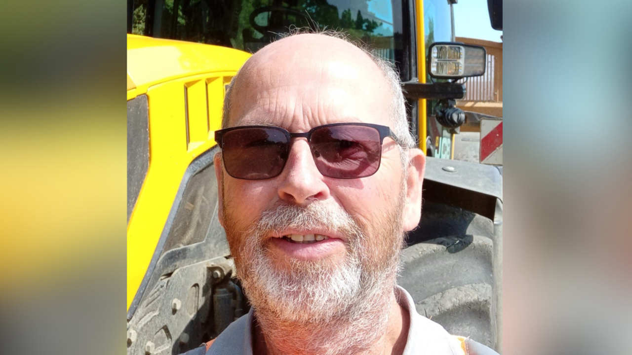 A selfie-style image of a man who is wearing dark glasses and standing in front of a tractor. He has a beard and is looking directly into the camera.