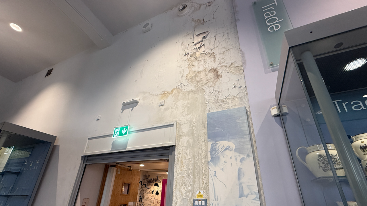 A wall damaged by water has peeling paint. It is above a door and next to a display cabinet. 