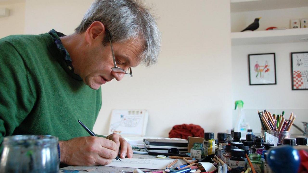 Axel Scheffler drawing in his studio. He is wearing glasses and is holding a pen over a drawing his desk where he is surrounded by pots of pens, inks and pencils