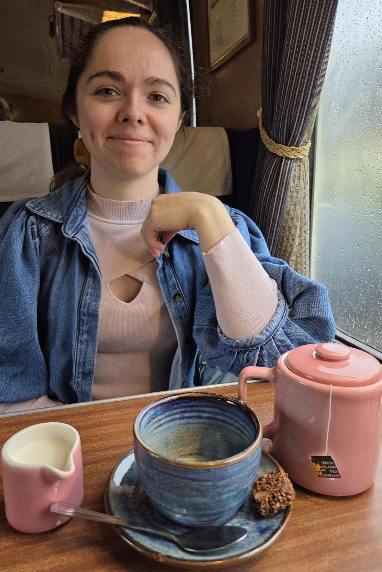 Rachael Robertson is sitting at a table next to a window. She has her dark hair tied back and is wearing a pink top under a blue denim shirt. On the table in front of her is a pink pot of tea, small pink jug of milk and a blue cup and saucer.