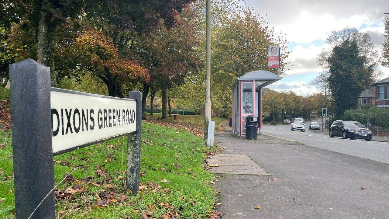 A side-on view of a road sign saying Dixons Green Road, which is situated on some grass. The grass is to the left of a pavement, which has a bus stop and lamppost, and a road with some cars on. Houses are just visible in the distance to the right-hand side of the road, while trees are on the grass on the left.