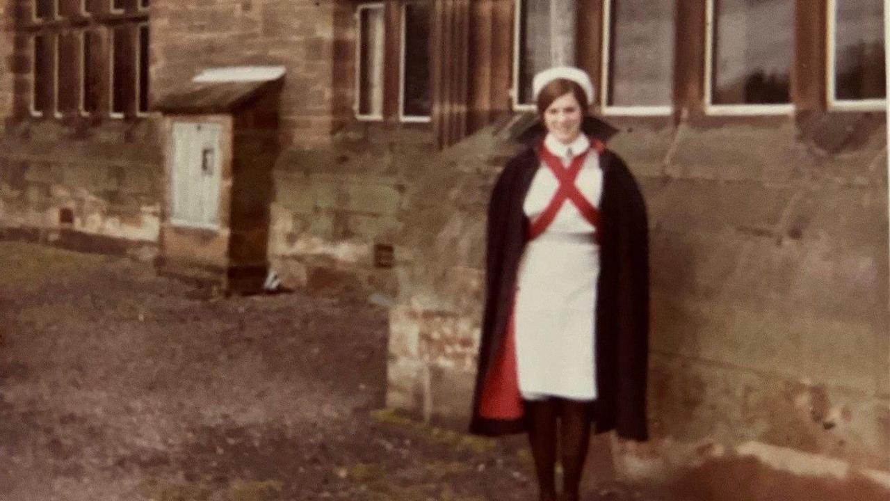 Beverley Pedley, in a white dress and black cape with red lining, is stood next to a large stone building which appears to be a hospital.