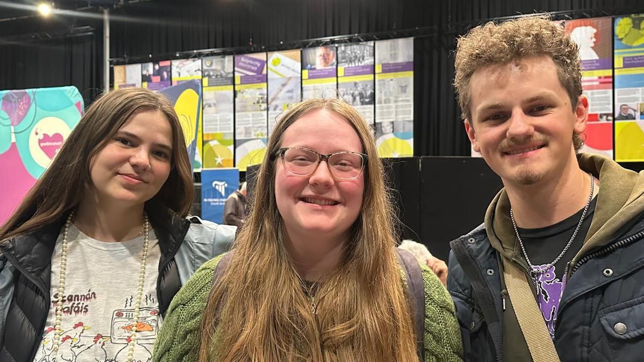 Three young people standing next to each other. Fiona has long dark brown hair and is wearing a padded jacket, and t-shirt with chickens on it. Preston has short light brown curly hair and a moustache and small beard. He is wearing a black padded jacket, light brown hoodie and black t-shirt.