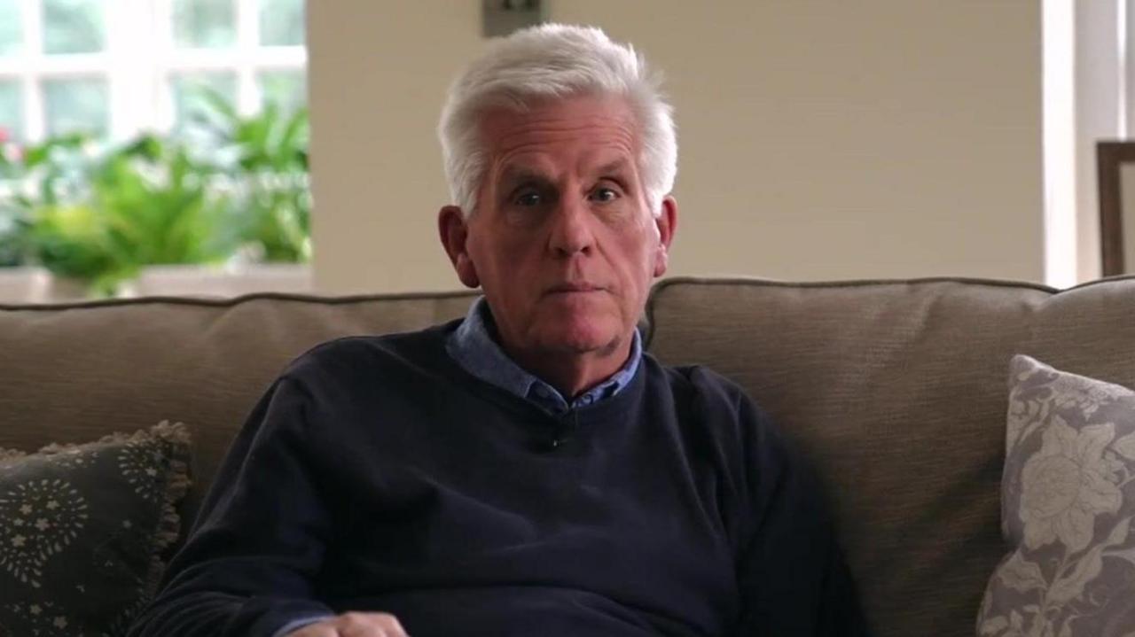 An elderly man looking at the camera. He has white hair and is wearing a navy jumper over a lighter blue collared shirt.